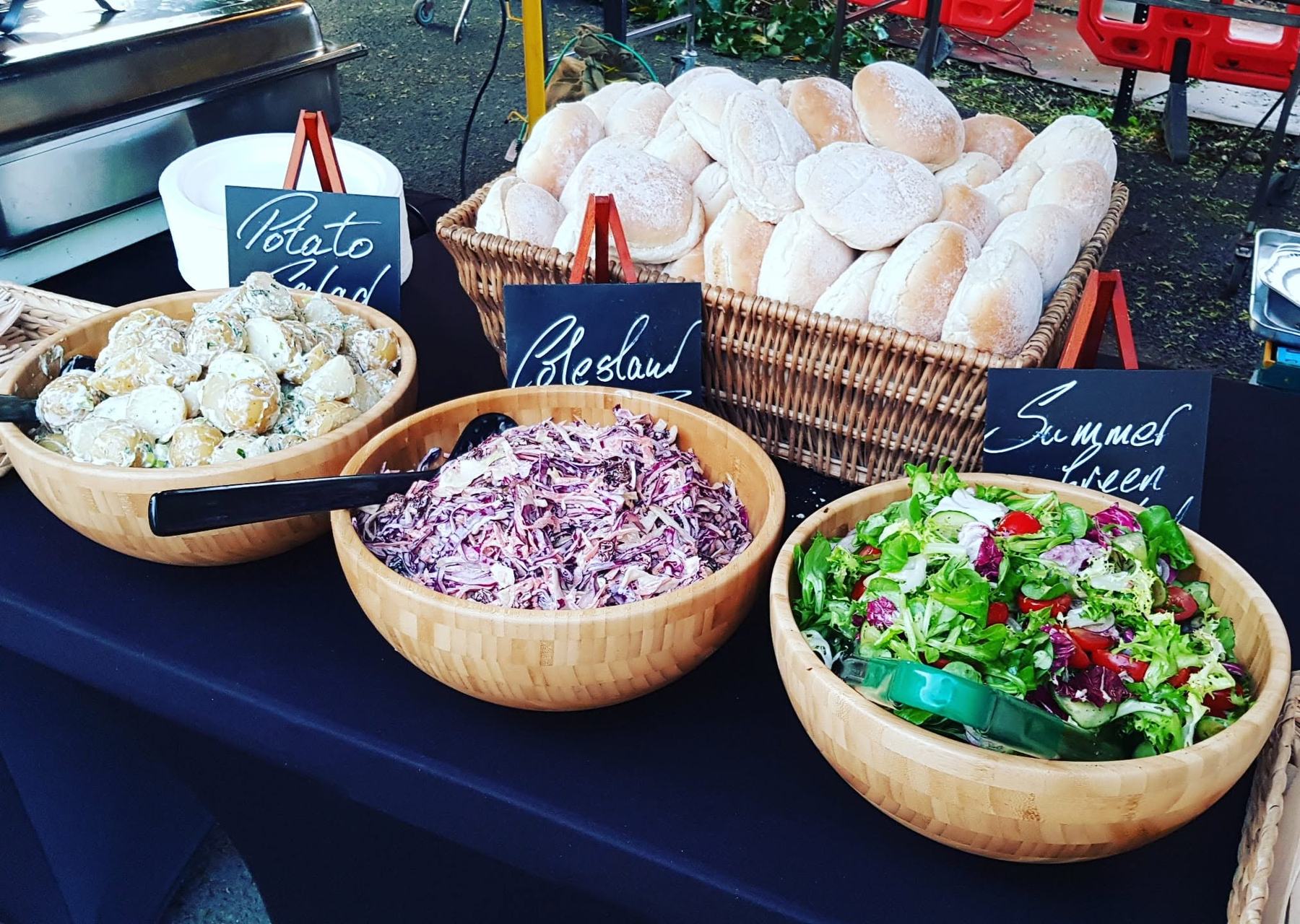 Bowls of potato salad, coleslaw and summer green salad with a basket of fresh rolls and chalkboard labels