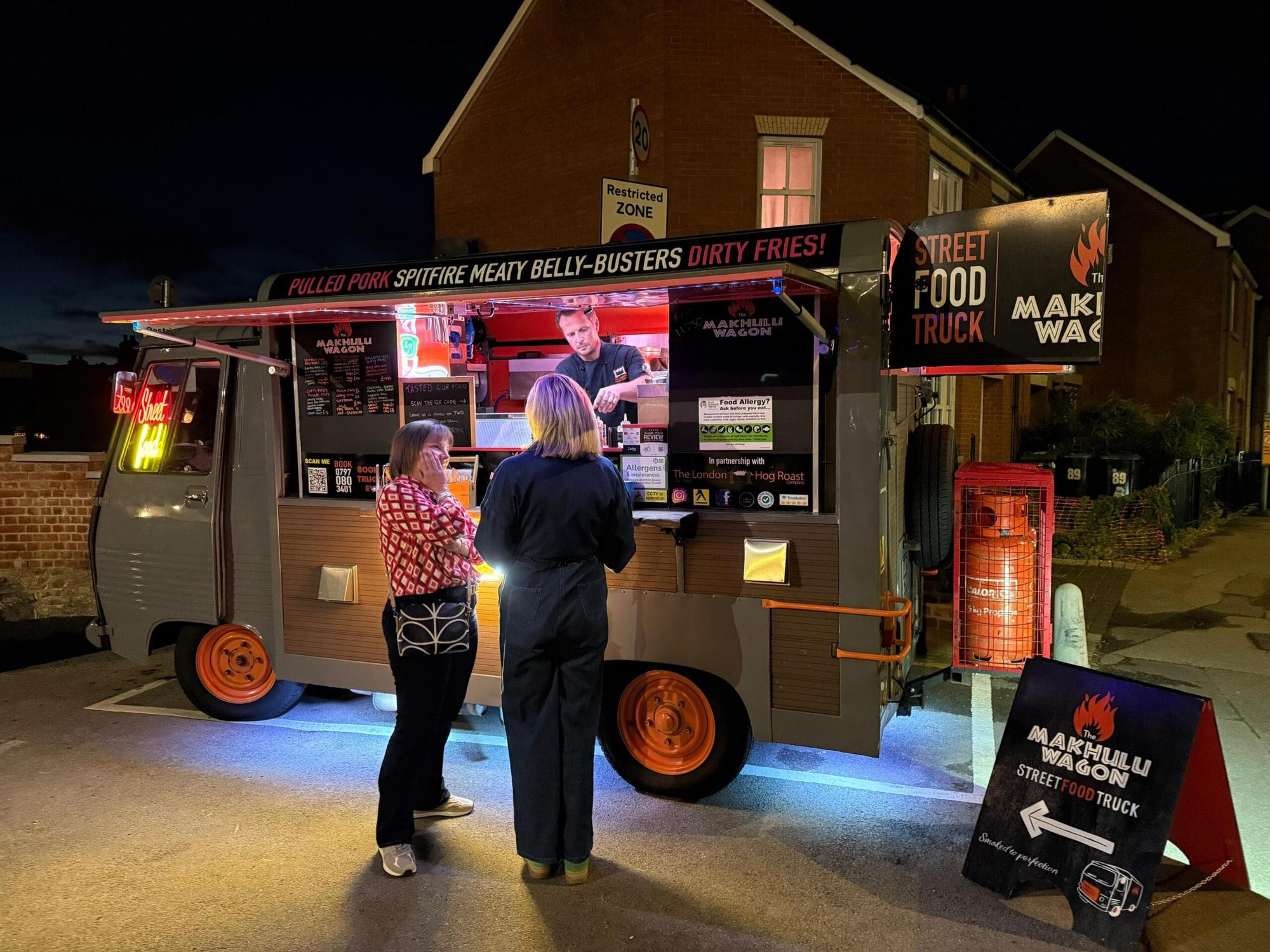 The Makhulu Wagon food truck lit up at night with customers ordering